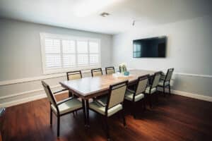 Conference room with large dining table, flat screen TV on the wall, hardwood floors and large window with white shutters.