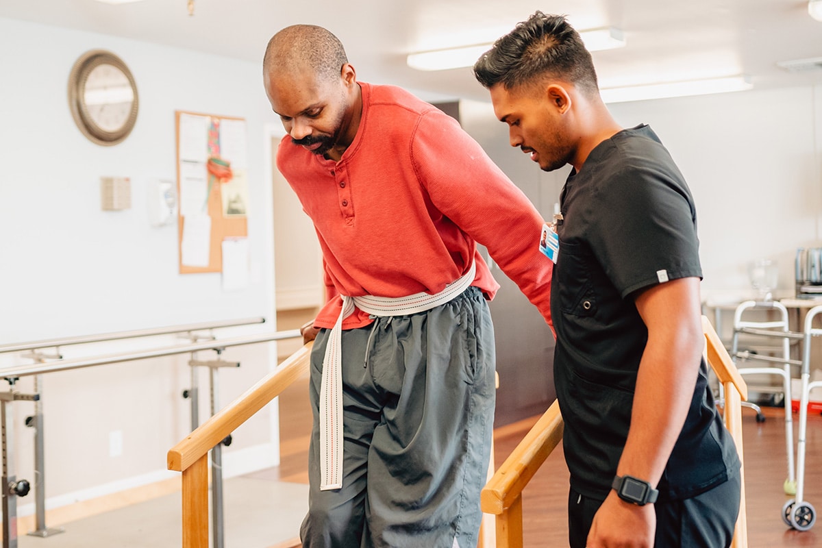 Physical therapist helping man in red shirt walk over a rehab bridge.