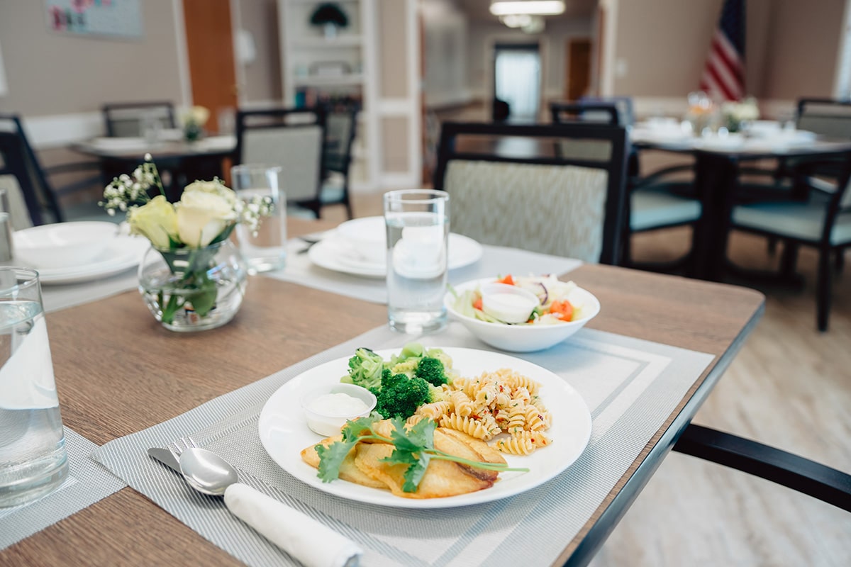Close up in dining room of a plate of fish, pasta, and brocoli with a side salad on a dining table with light green placemats and roses on the table.
