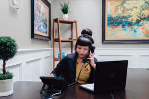 Woman with black hair in bun sitting at reception desk answering phone and laptop open on her desk.
