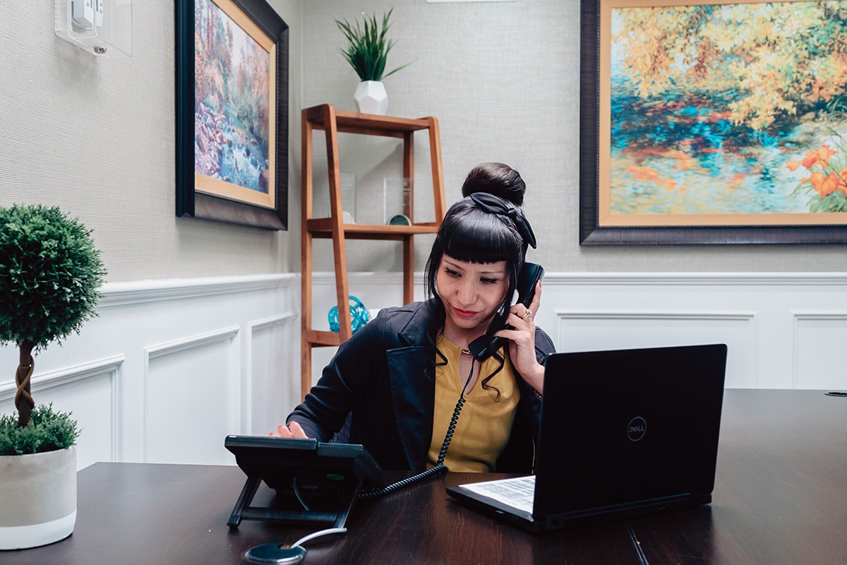 Woman with black hair in bun sitting at reception desk answering phone and laptop open on her desk.